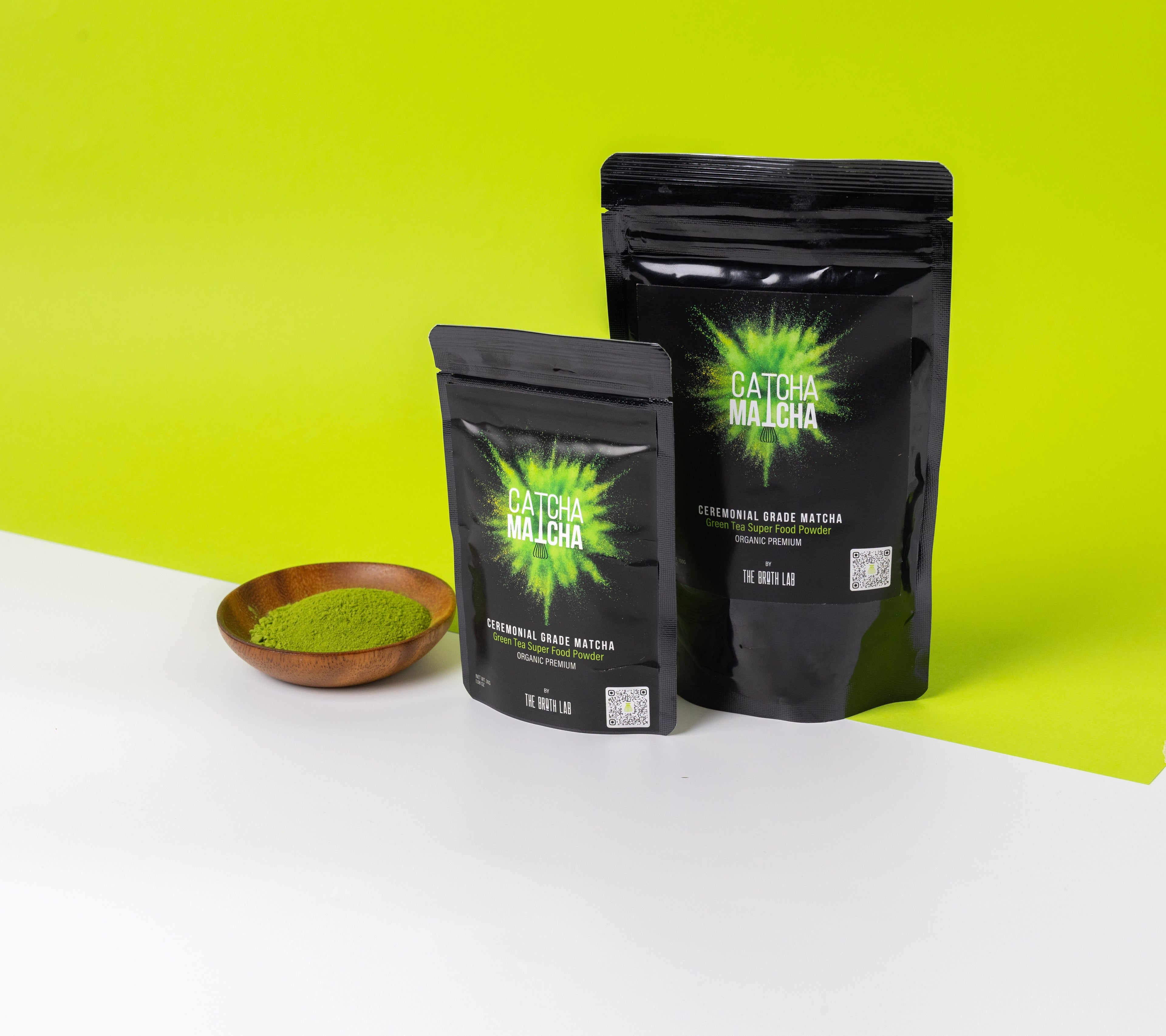 Two black packages of 'Catcha Matcha' matcha powder on a white surface with a green background.