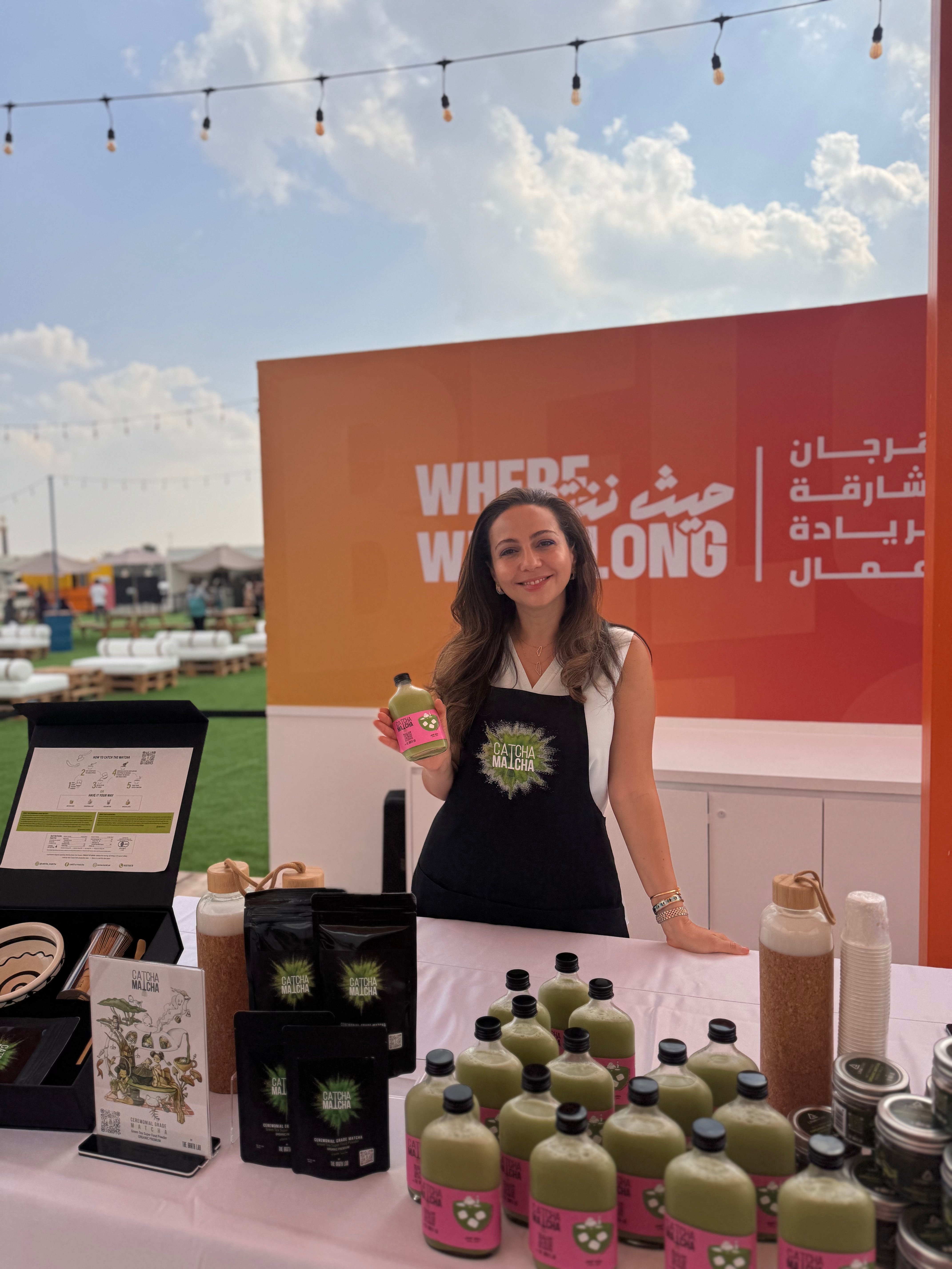 Hadil alkhatib holding a bottle of catcha matcha at a promotional event with branded backdrop in Sharjah