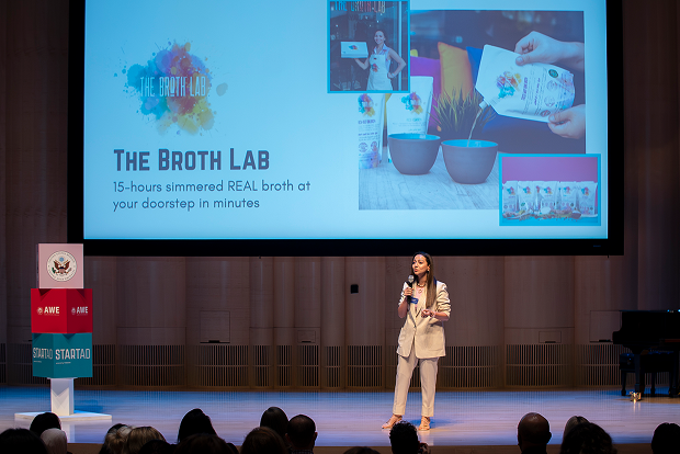 Woman giving a presentation on stage with a screen displaying 'The Broth Lab' in the background.